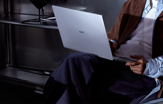 어두운 실내 공간에서 갈색 재킷을 입은 사람이 무릎 위에 은색 LG 그램 노트북을 올려두고 사용하고 있다. 노트북 상단에는 ‘gram’ 로고가 보이며, 배경에는 책상과 스탠드 조명이 있다